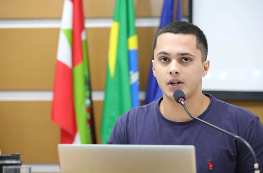 A imagem retrata um homem jovem de pele clara e cabelos castanhos curtos, falando ao microfone em um ambiente que aparenta ser uma câmara legislativa ou auditório oficial.