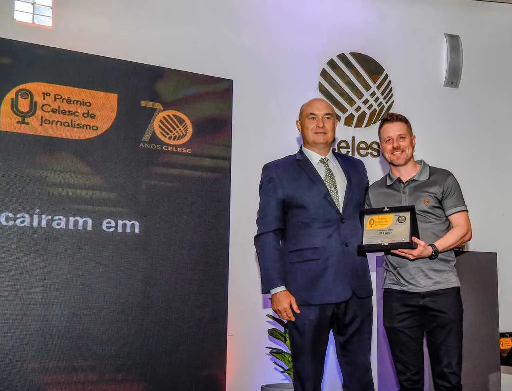 A imagem mostra duas pessoas em um palco ou evento de premiação, posando para uma foto. Um dos homens está entregando uma placa de reconhecimento ao outro.